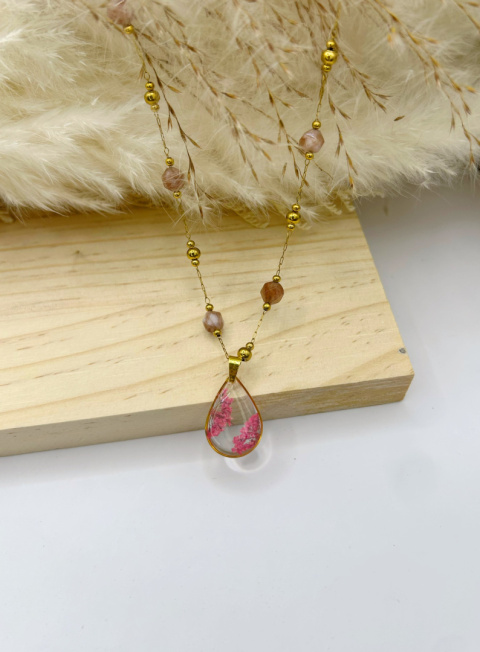Naszyjnik handmade – przezroczysta, kroplowa zawieszka z żywicy z zatopionymi różowymi suszonymi kwiatami na pozłacanym łańcuszku zdobionym fasetowanymi kamieniami słonecznymi, na miękkim, beżowym tle.