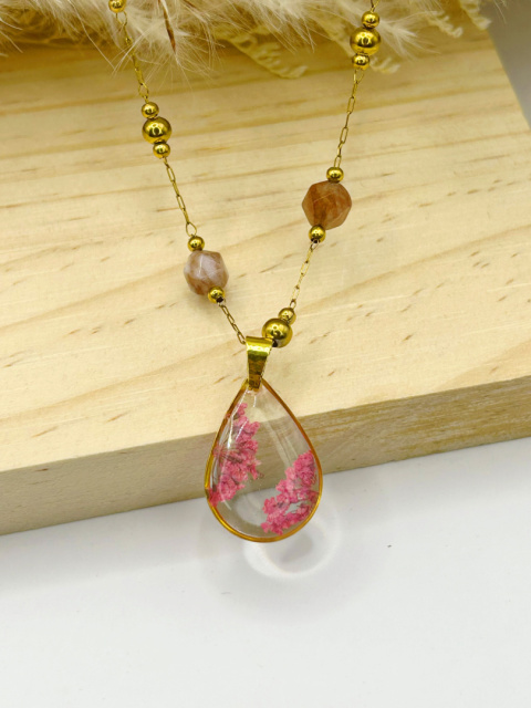 Naszyjnik handmade – przezroczysta, kroplowa zawieszka z żywicy z zatopionymi różowymi suszonymi kwiatami na pozłacanym łańcuszku zdobionym fasetowanymi kamieniami słonecznymi, na miękkim, beżowym tle.