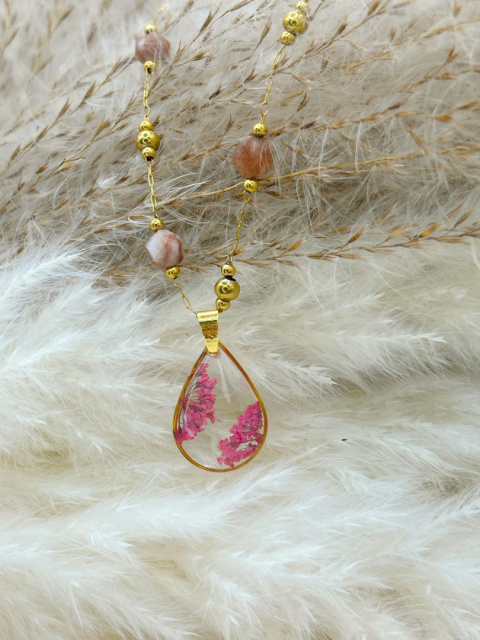 Naszyjnik handmade – przezroczysta, kroplowa zawieszka z żywicy z zatopionymi różowymi suszonymi kwiatami na pozłacanym łańcuszku zdobionym fasetowanymi kamieniami słonecznymi, na miękkim, beżowym tle.